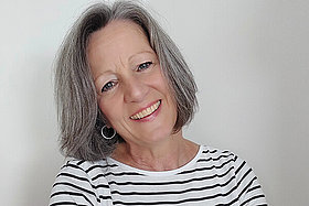 Frau um die 60, schulterlange dunkle, grau melierte Haare und schwarz-weiß gestreiftes Shirt. 