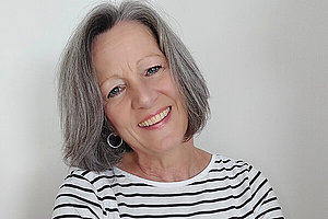 Frau um die 60, schulterlange dunkle, grau melierte Haare und schwarz-weiß gestreiftes Shirt. 