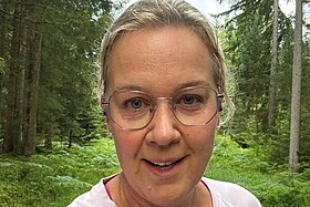 Blonde Frau mit Brille steht im Wald und schaut lächelnd in die Kamera