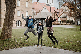 Vor einem Bauernhaus: eine Familie (Mutter, Vater, Kind (Junge, etwa 11 Jahre alt) tragen Wintersachen und springen fröhlich in die Luft. 
