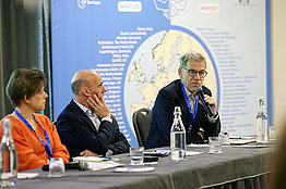 Blick auf ein Podium bei einer Konferenz, da sitzen eine Frau und zwei Männer, einer der Männer spricht gerade.