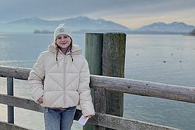 Junge Frau mit weißer Mütze und Zöpfen steht vor einem Holzgeländer an einem See - im Hintergrund sieht man Berge. 