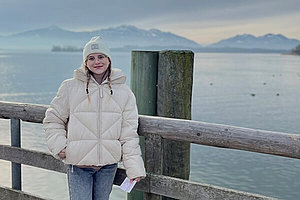 Junge Frau mit weißer Mütze und Zöpfen steht vor einem Holzgeländer an einem See - im Hintergrund sieht man Berge. 