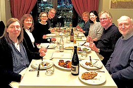 Acht Personen sitzen scheinbar in einem Restaurant an einem längeren Tisch und schauen in die Kamera. 