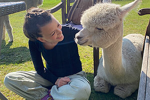 Eine Frau im dunklen Shirt sitzt auf einer Wiese im Sonnenschein neben einem weißen Alpaka und streichelt es. 