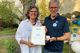 Susi Pfeiffer-Auler erhielt die Adolf-Windorfer-Medaille von Stephan Kruip. (Sie stehen im Garten und halten gemeinsam Urkunde und Medaille in die Kamera).