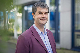 Porträt eines Mannes um die 50 mit weinrotem Jackett, im Hintergrund sieht man einen Baum. 