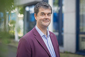 Porträt eines Mannes um die 50 mit weinrotem Jackett, im Hintergrund sieht man einen Baum. 