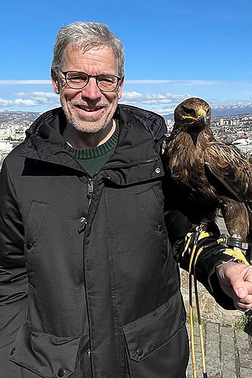Mann - um die 60 Jahre alt - hat einen Falken auf dem Arm (inklusive Schutzkleidung am Arm), der Himmel im Hintergrund ist blau. 