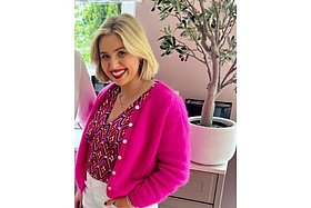 Junge Frau mit halblangen blonden Haaren und pinker fluffiger Strickjacke, im Hintergrund sieht man eine Zimmerpflanze. 
