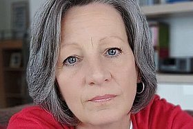 Porträt von Simona, graue Haare, rote Strickjacke, ca. 55 Jahre alt. Sie blickt in die Kamera. 