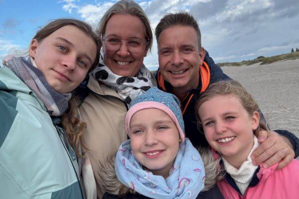 Mutter, Vater und drei Mädchen mit Windjacken stehen am Meer und lächeln in die Kamera