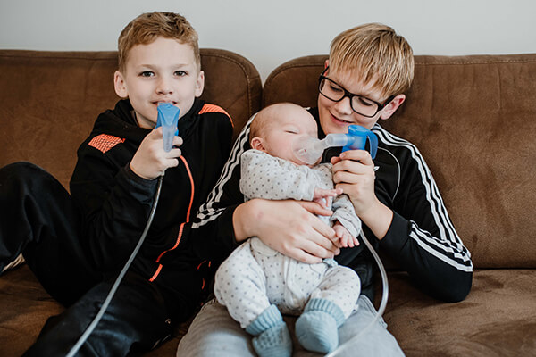 Drei Jungen sitzen auf einem Sofa, der ältere hält seinen kleinen Bruder, ein Baby auf dem Schoß, und hilft ihm beim Inhalieren. Auch der mittlere Junge inhaliert. 