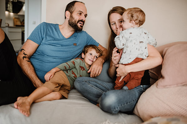 Emil mit seiner Familie Eine Familie (Vater, Mutter und zwei kleine Kinder) sitzen auf einem Sofa. Sie lachen.