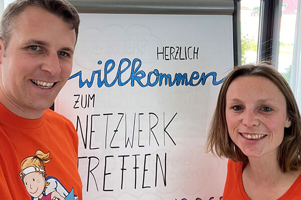 Erstes Netzwerktreffen in der Region Fulda Mann und Frau in orangenen T-Shirts stehen vor einem Flipchart, auf dem man "Herzlich Willkommen" und "Netzwerktreffen" lesen kann.