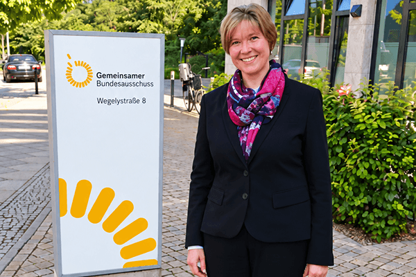 Eine Frau mit kurzen blonden Haaren und Anzug steht vorm Eingangsschild des Gemeinsamen Bundesausschusses (G-BA)