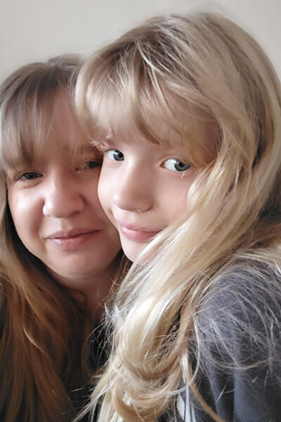 Mutter und Tochter, beide mit leicht gelockten blonden Haaren und Pony, blicken in die Kamera. Man sieht eine Ähnlichkeit zwischen den beiden. 