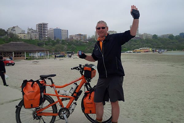 Mann steht mit einem Fahrrad an einem Strand, der nicht in Deutschland ist. Der Arm, eher mittelalt, reckt einen Arm in die Höhe. 
