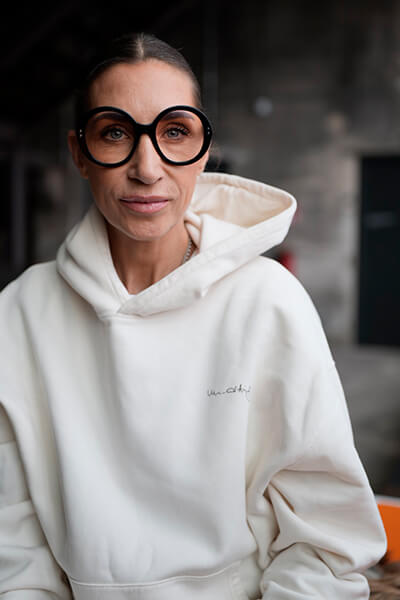 Frau mit weißem Kapuzenpulli, zusammengebundenen Haaren und dick umrandeter Brille