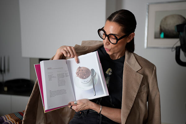 Frau mit dunklen Haaren, dick umrandeter Brille hält ein aufgeschlagenes Buch in die Kamera, darin erkennt man ein Rezept für Eiscreme