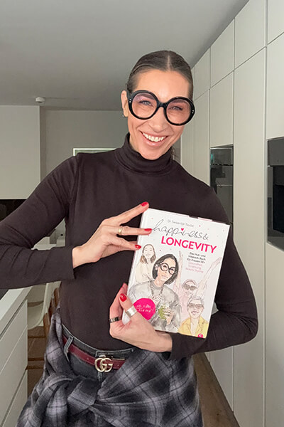 Frau mit dick umrahmter Brille und braunem Rollkragenpullover hält das Buch „Happiness & Longevity – Das Mutmachbuch für Frauen 50+“ in die Kamera. 