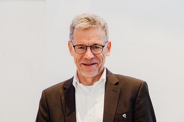 Porträt eines Mannes um die 60 Jahre mit Hemd und Anzug, er trägt eine Brille. 